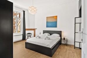A modern bedroom with a large bed, a view of buildings through a window, a painting on the wall, and a hanging light fixture.