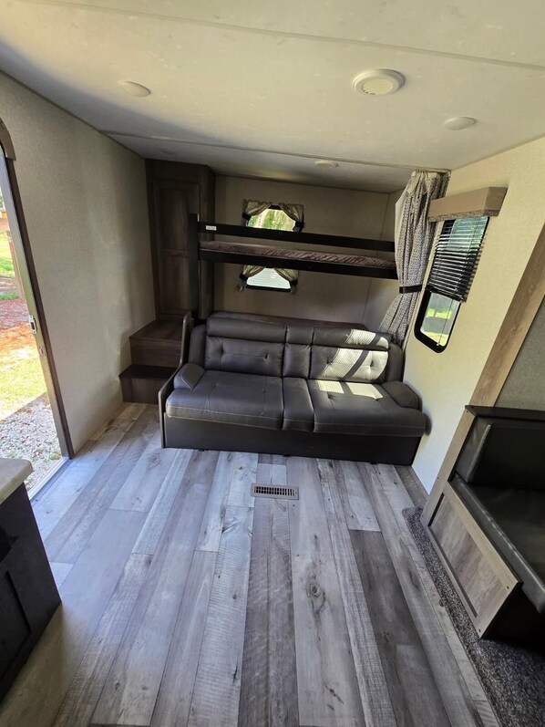 A compact RV interior with a sofa bed, a window with curtains, and a small kitchenette area.