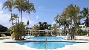 Un área de piscina con palmeras, un espacio de descanso cubierto y un cielo azul despejado.