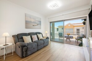 A modern living room with a grey sofa, a small table with a lamp, and a sliding glass door leading to an outdoor patio. - Apartment 'T2 Reis' with Shared Pool, Wi-Fi, and Air Conditioning (Albufeira)