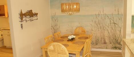 A dining area with a wooden table, wicker chairs, and a wall mural of a beach scene.