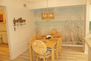 Un comedor con mesa de madera, sillas de mimbre y un mural en la pared que representa una playa.