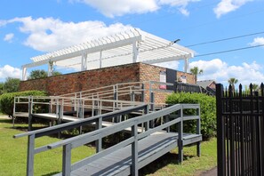 A brick building with a white pergola, a gated entrance, and a wooden ramp leading to a covered area.