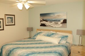 Un dormitorio con una cama, un ventilador de techo, un cuadro de una playa y dos lámparas de noche.