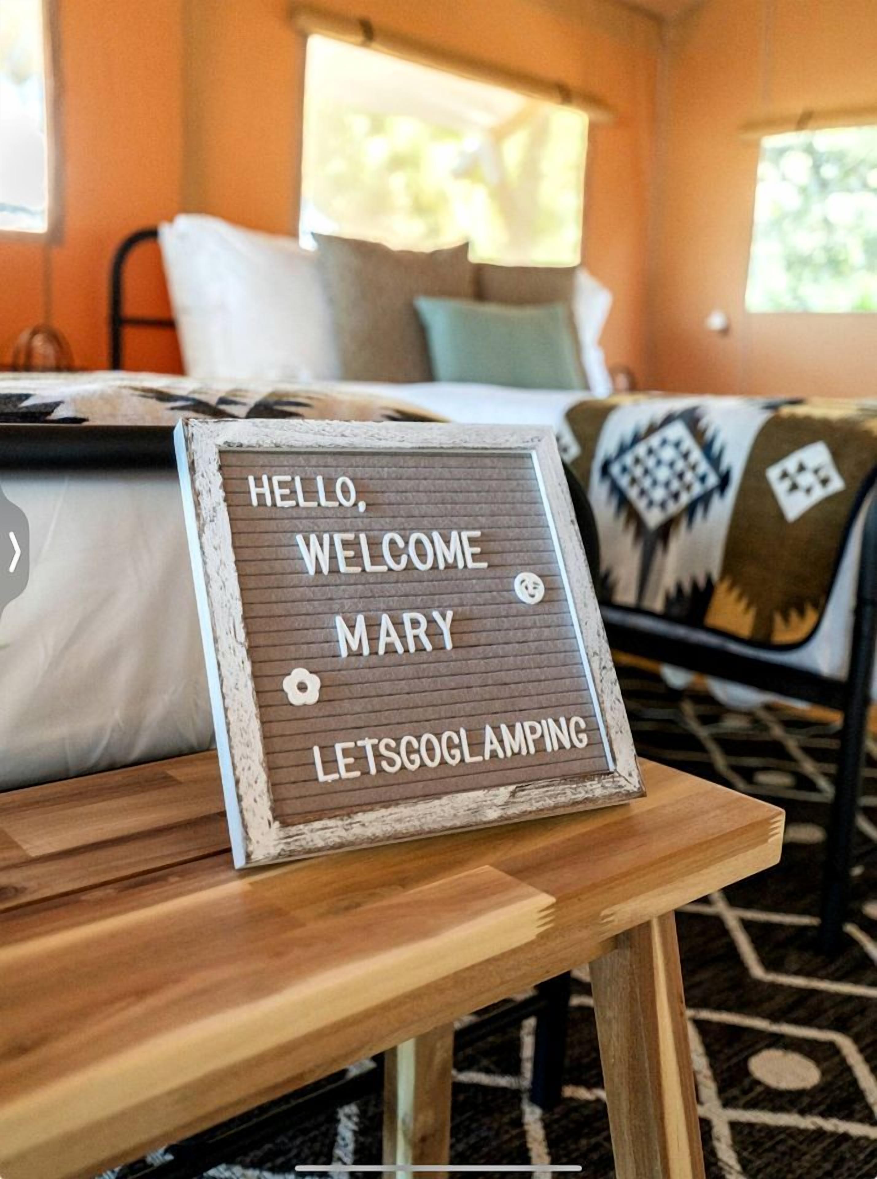 A bed with a sign that reads 'HELLO, WELCOME MARY LETSGOGLAMPING' on a wooden table.