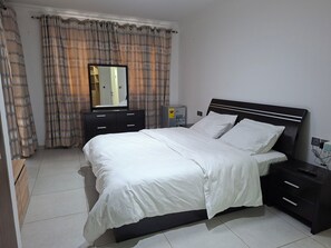 A neatly made bed with white linens, a dark headboard, and a nightstand with a remote control. - Cozy 1-bedroom apartment with a pool, standby genset in tranquil West Hills (Accra)