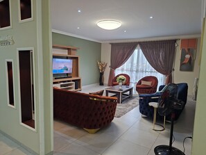 A living room with a TV, red and blue sofas, a table, and a standing fan. - Cozy 1-bedroom apartment with a pool, standby genset in tranquil West Hills (Accra)