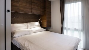 Una camera d'albergo moderna con un letto grande, una testiera in legno e una finestra con tende trasparenti.