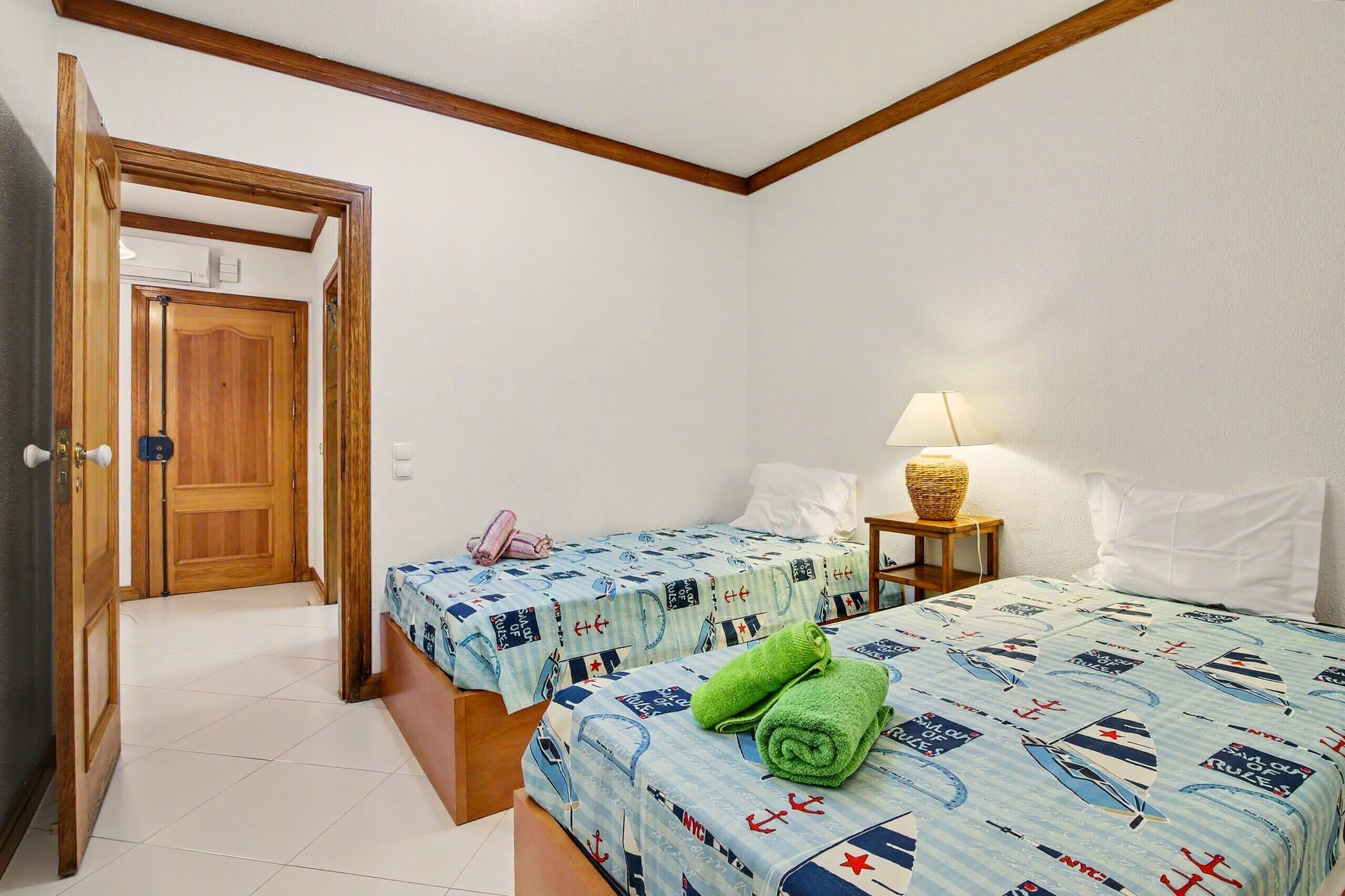 A room with two beds, a wooden door, a lamp, and a small table.