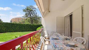A balcony with a red railing, a table set with plates and glasses, and a view of a building and greenery.