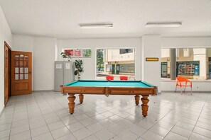 Ein Billardtisch in einem Raum mit weiß gefliestem Boden, einer Holztür und einem Fenster mit Blick auf die Straße.