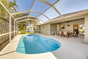 Ein überdachter Pool mit Glasdach, eine Terrassenfläche mit Stühlen und Tisch sowie Blick auf Palmen.