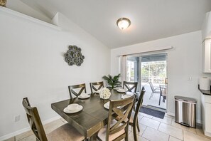 Ein Essbereich mit Tisch für vier Personen, Stühlen, einer Wanddekoration und Blick durch eine Glastür auf eine Terrass.'