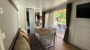 A room with a sofa, a dining table with chairs, a television on the wall, and a view of an outdoor patio. - Terme Tuhelj Glamping Village: Vita Family Mobile Home for 6 People (Tuheljske Toplice)