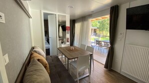 A room with a sofa, a dining table with chairs, a television on the wall, and a view of an outdoor patio. - Terme Tuhelj Glamping Village: Vita Family Mobile Home for 6 People (Tuheljske Toplice)