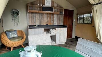 A tented accommodation with a green floor, a white lantern, a wicker chair, and a wooden shelf with various items.