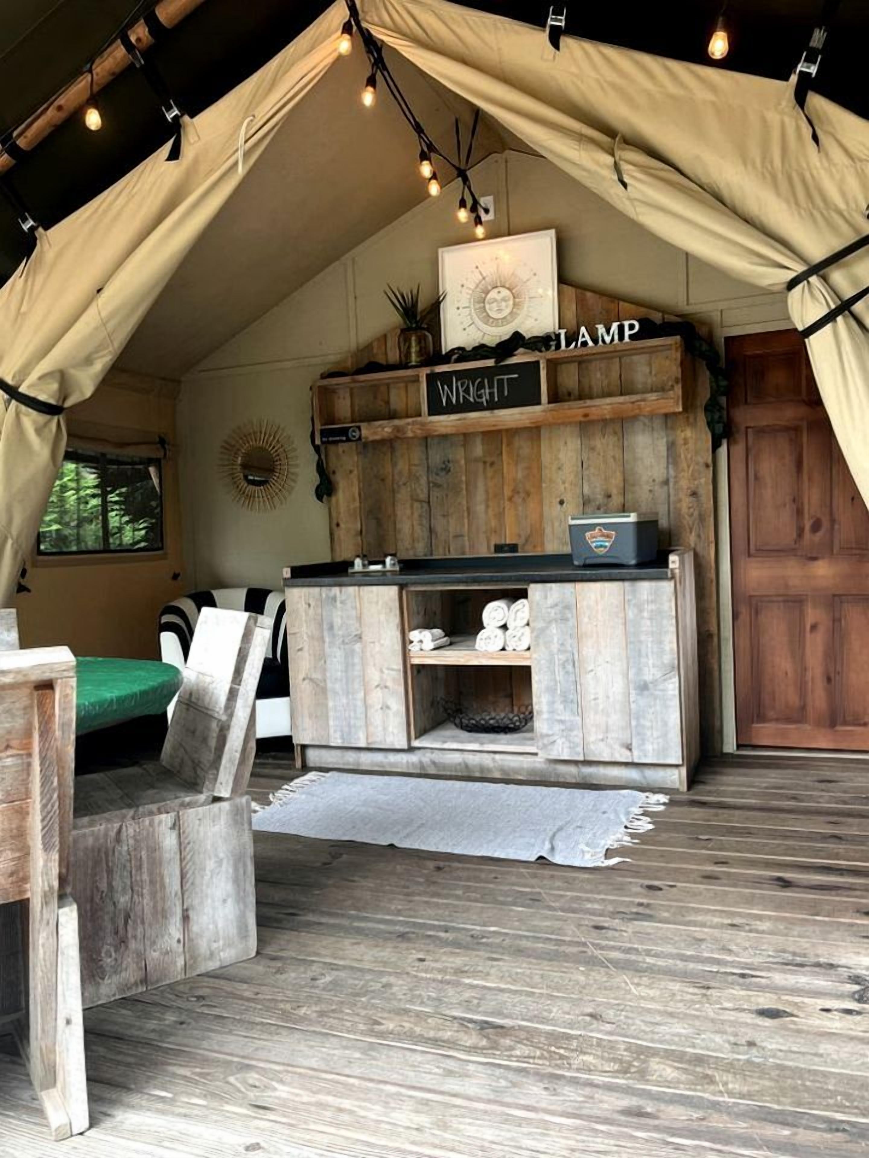 A rustic tent interior with a wooden bench, a sign reading 'Wright Lamp,' and a small table with a lamp.