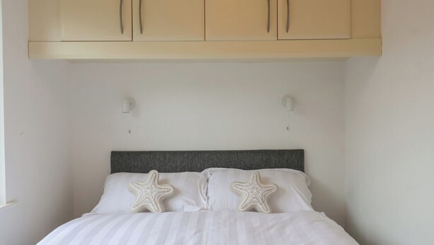 A bed with a striped white duvet, two starfish pillows, a grey headboard, and beige cabinets above. - Lodge 19 (Torpoint)
