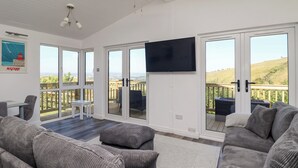 A spacious living room with a large TV, grey upholstered sofas, a ceiling fan, and a view of the countryside through large sliding doors. - Lodge 19 (Torpoint)