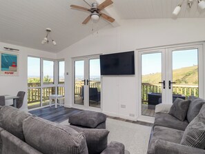 A spacious living room with a large TV, grey upholstered sofas, a ceiling fan, and a view of the countryside through large sliding doors. - Lodge 19 (Torpoint)