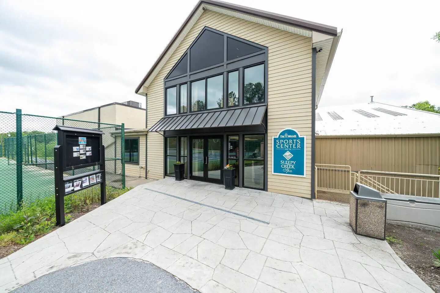 Ein Sportzentrum mit großem Glaszugang, einem Schild, das es als Teil von The Woods kennzeichnet, und einem eingezäunten Tennisplatz.