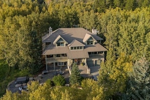 A large, two-story house surrounded by dense forest, with a prominent front porch and multiple windows.