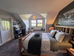 A bedroom with a bed, a bench, a TV, and a window with a view. - Queen Bdrm | Private Bthrm | Balcony | Bragg Creek (Bragg Creek)