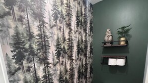 Une salle de bain avec un papier peint à motifs de forêt, des toilettes, une poubelle et une étagère sur laquelle se trouvent une plante et un objet décoratif.