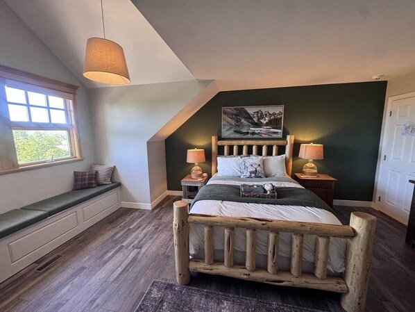 A bedroom with a sloped ceiling, a wooden bed frame, a bench by the window, and a painting on the wall.