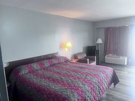A hotel room with a large bed, a desk, a chair, a television, and an air conditioning unit.