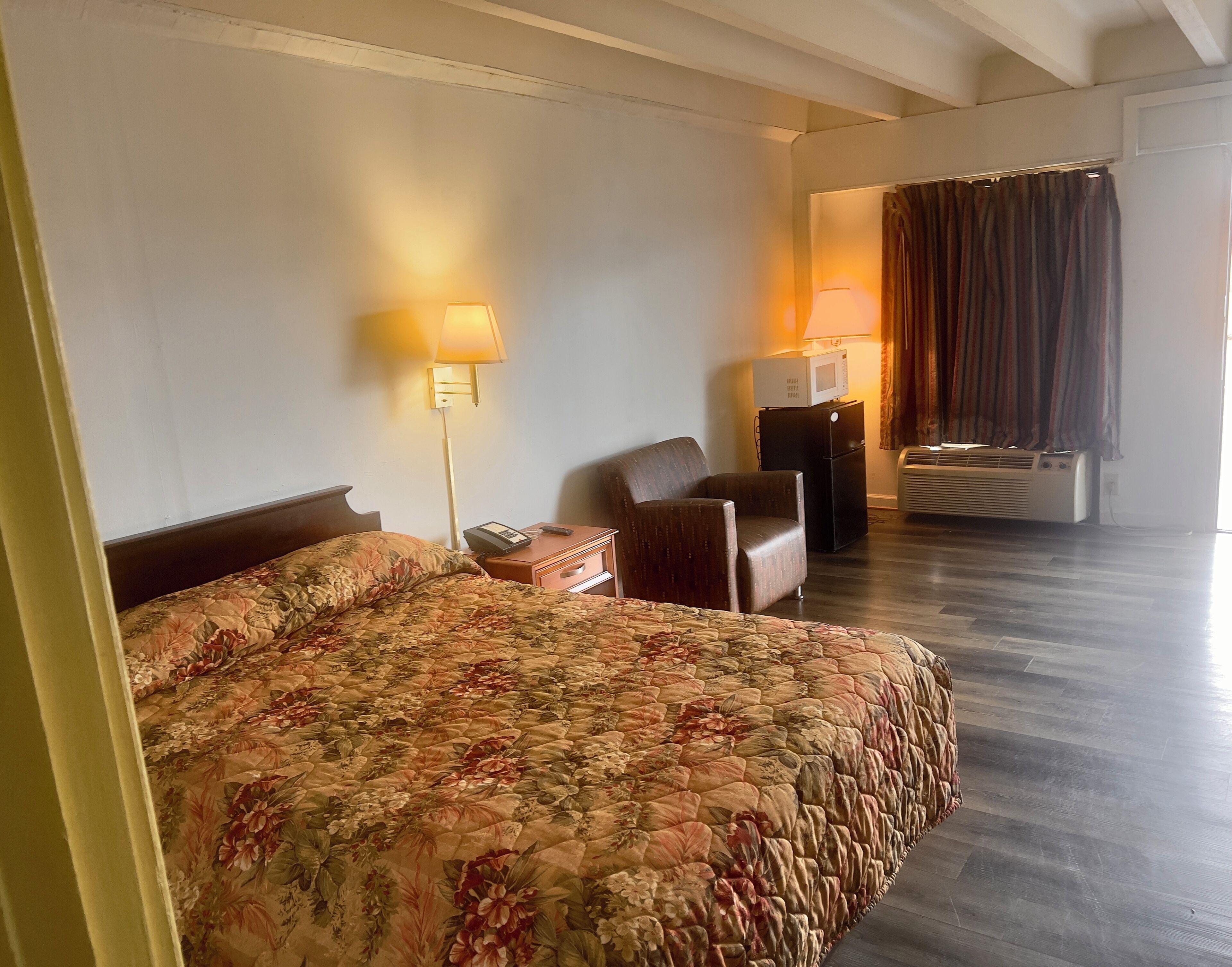 Quarto de hotel com uma cama grande, uma escrivaninha, uma cadeira, uma luminária, um micro-ondas e um aparelho de ar condicionado.