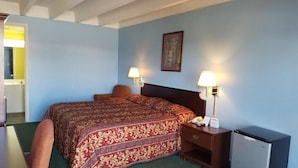 Habitación de hotel con cama, lámparas de noche, un escritorio pequeño con lámpara, refrigerador y un cuadro en la pared.