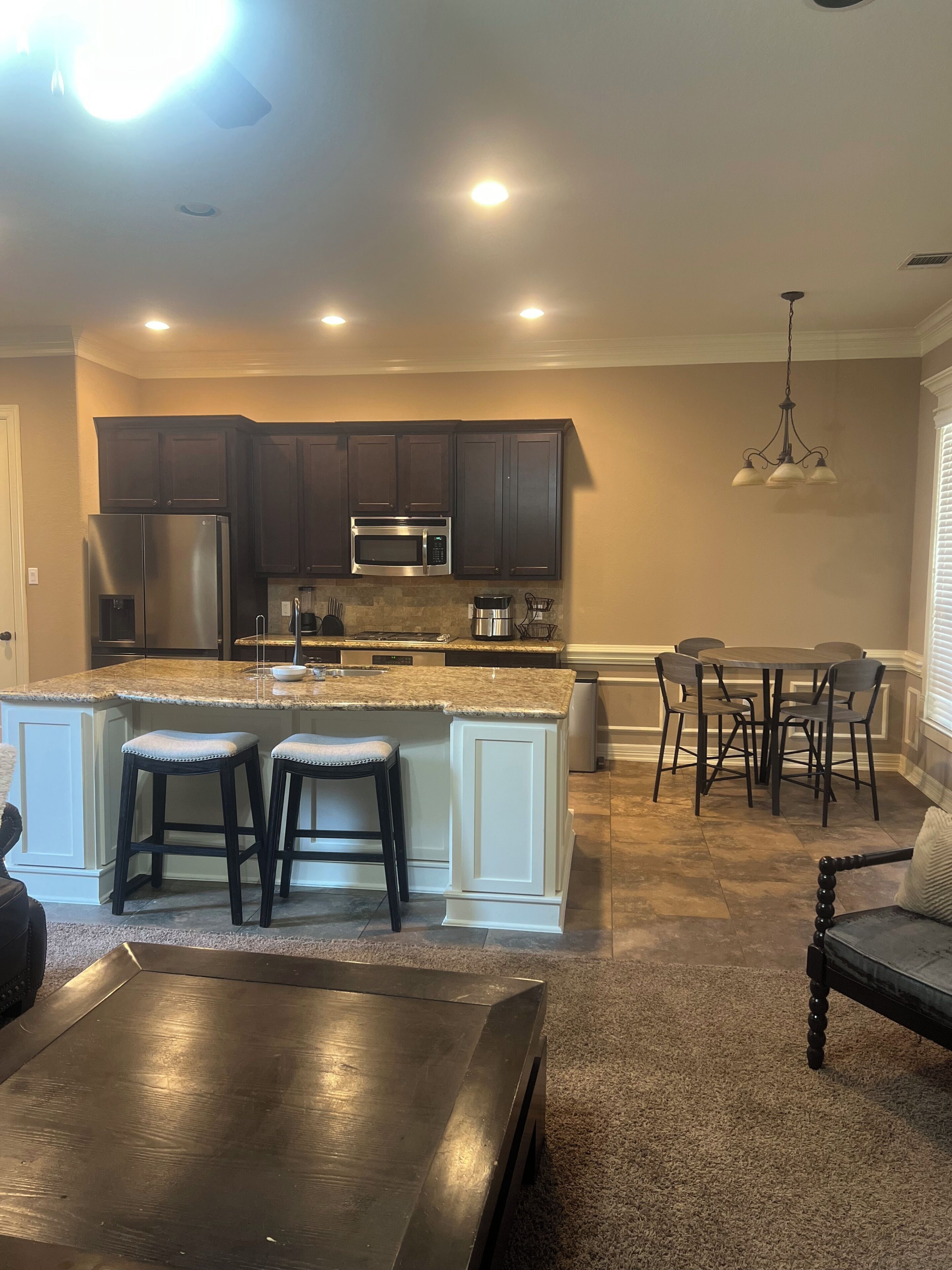 A kitchen with dark cabinets, a stainless steel refrigerator, a microwave, and a three-burner stove. There is a bar area with stools and a dining table with chairs.