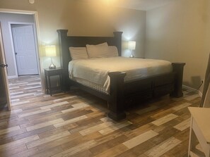 A bedroom with a large bed, two bedside tables with lamps, a wooden floor, and a door in the background. - Welcome to the Round Roost- 3 story+sleeps 16+Lake Ham+grill+decks+game room (Hot Springs)