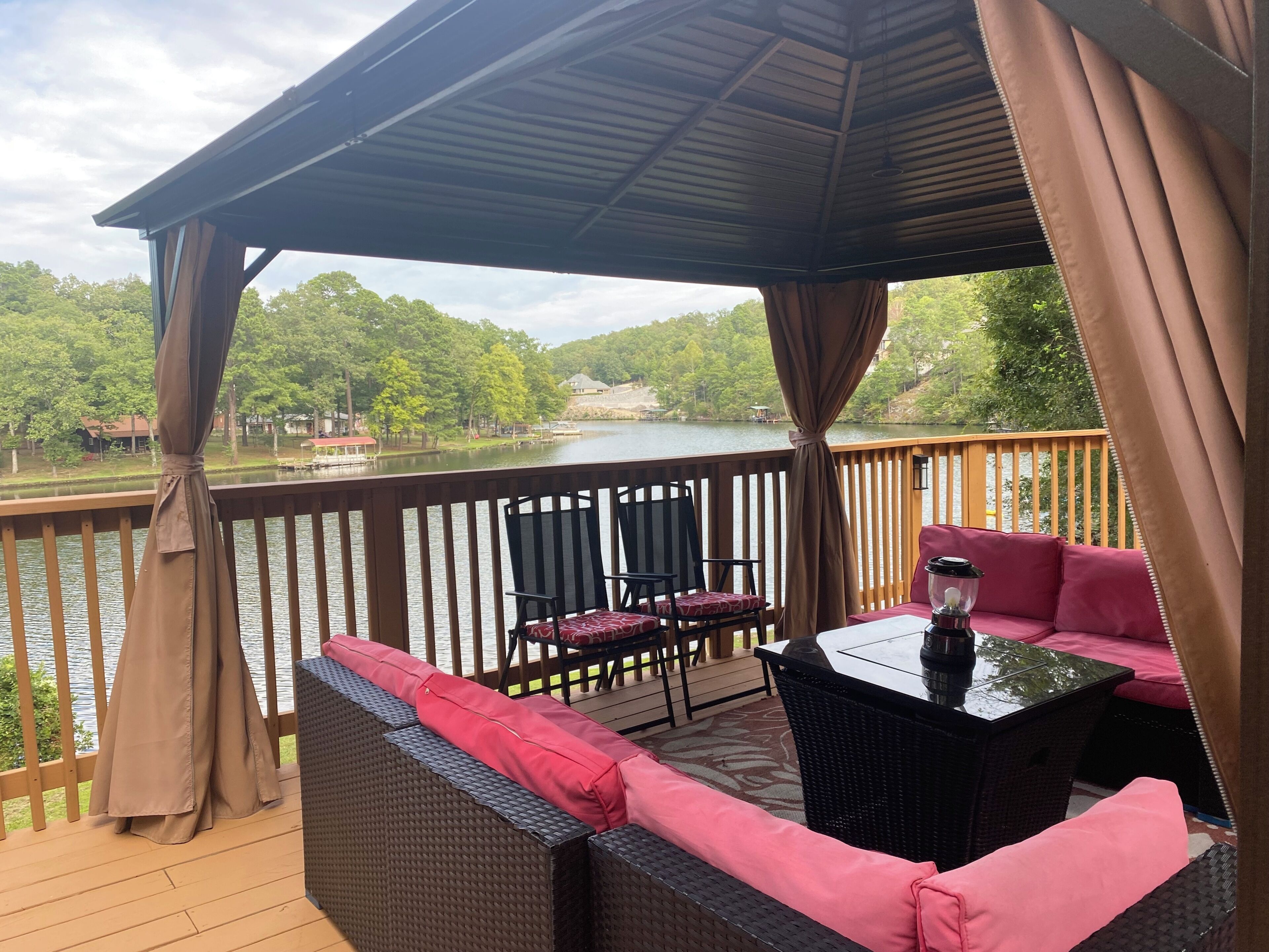Una terraza cubierta al aire libre con muebles de mimbre y una mesa, con vista a un lago con árboles y edificios al fondo.