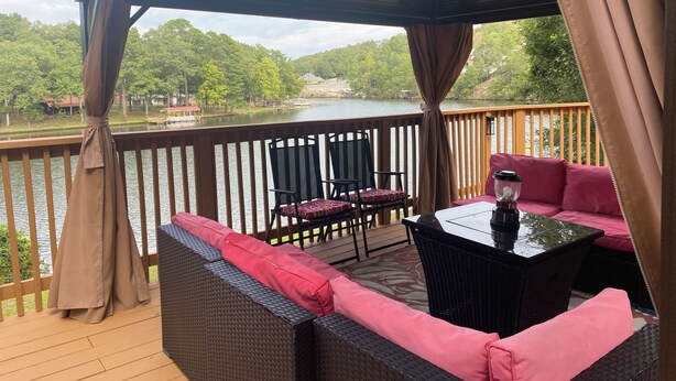 A covered outdoor deck with wicker furniture and a table, overlooking a lake with trees and buildings in the background. - Welcome to the Round Roost- 3 story+sleeps 16+Lake Ham+grill+decks+game room (Hot Springs)