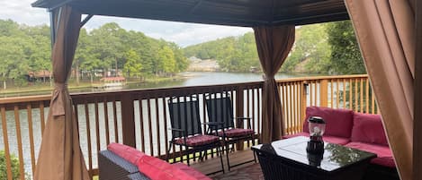 A covered outdoor deck with wicker furniture and a table, overlooking a lake with trees and buildings in the background.