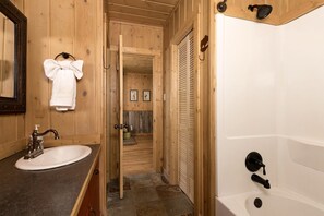Ein Badezimmer aus Holz mit Waschbecken, Spiegel, Handtuchhalter, Dusche und einer Tür zu einem Flur mit gerahmten Bildern.