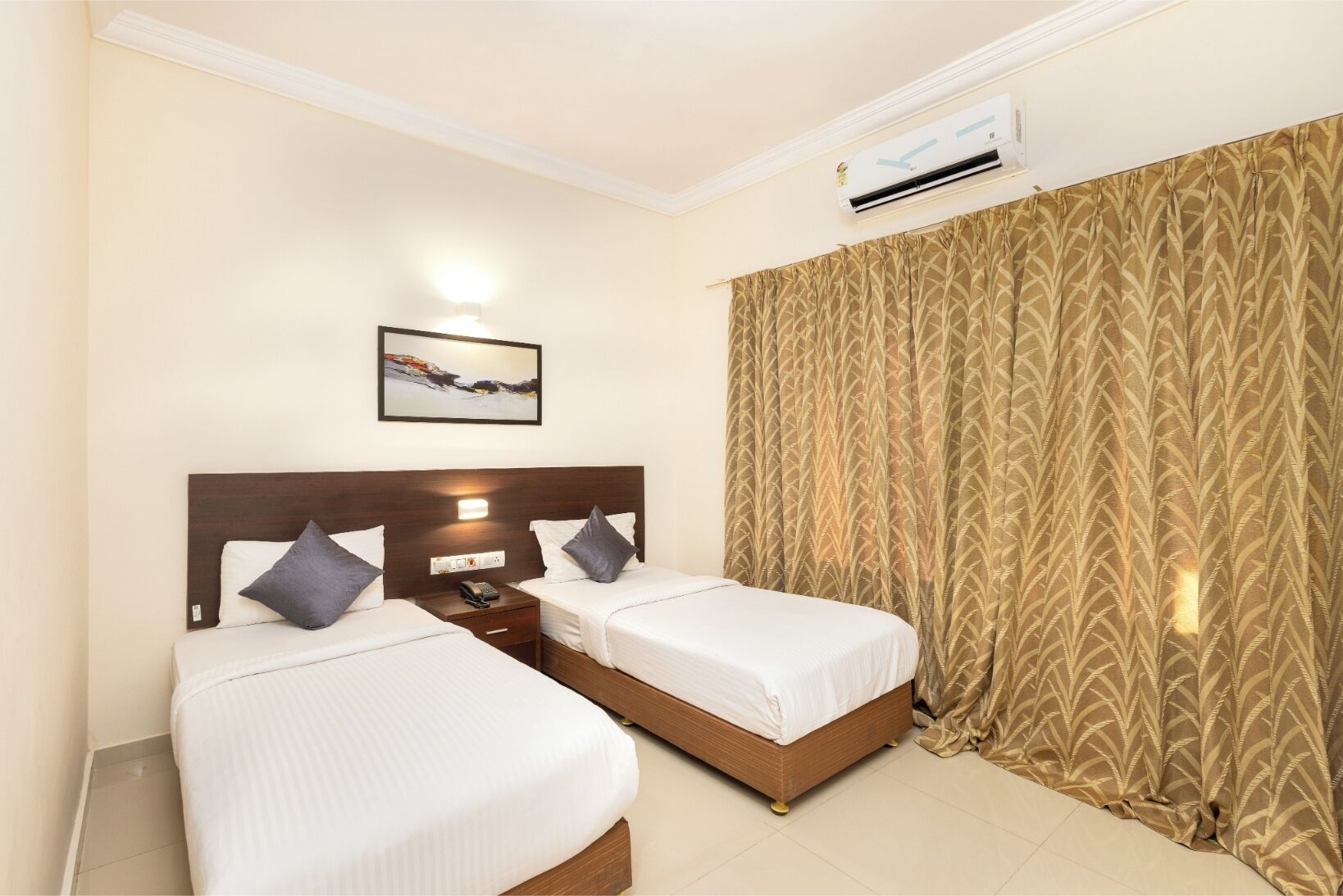 A hotel room with two beds, a nightstand, a wall-mounted air conditioner, a painting, and a patterned curtain.