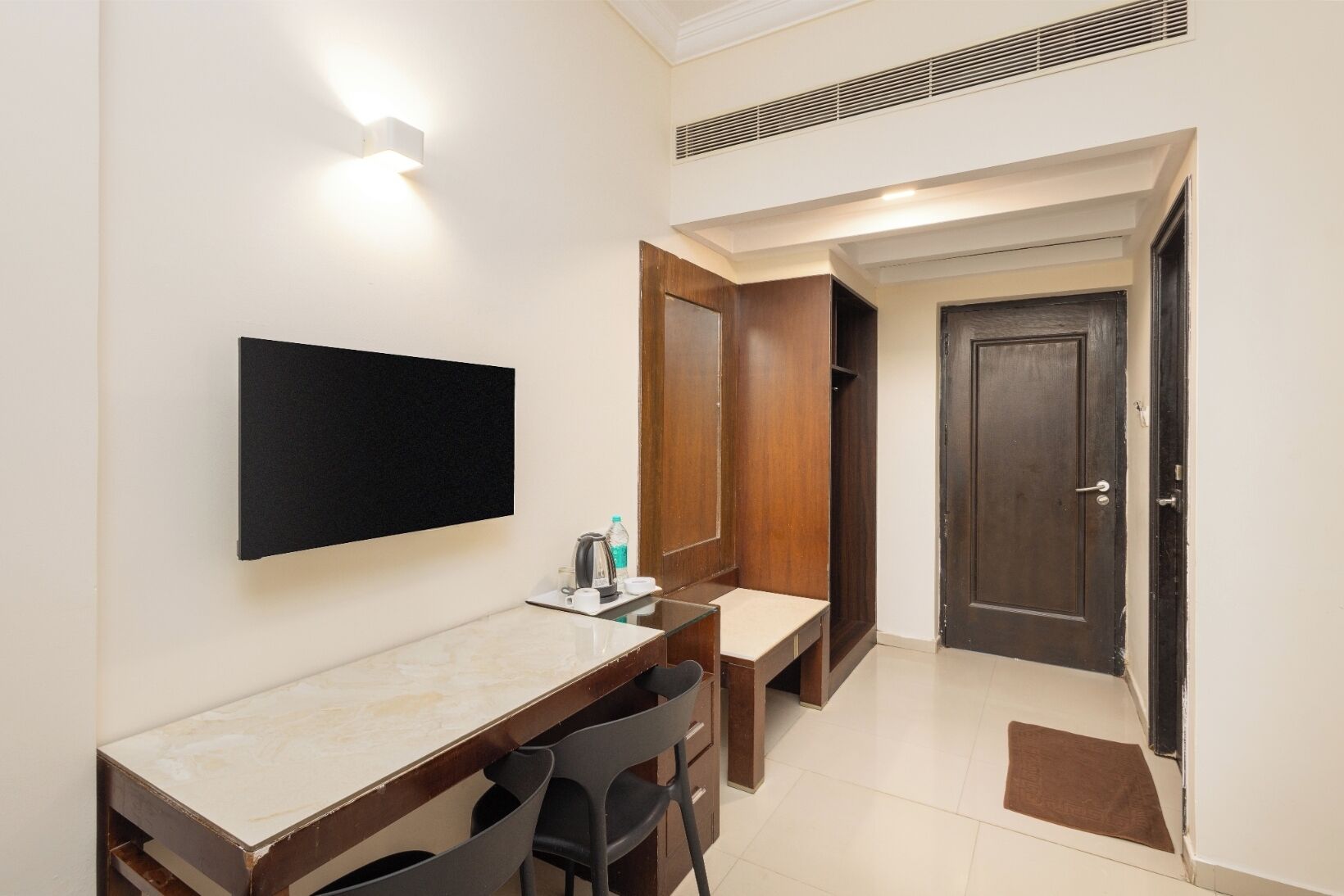 Habitación de hotel con televisor de pantalla plana montado en la pared, un escritorio de madera con sillas, una mesita con hervidor y tazas, y un armario con espejo.