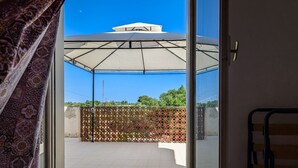 A view through a glass door to an outdoor patio area with a retractable awning, a patterned fence, and a clear blue sky. - The Sicilian Farmhouse (Palmeri - Ferrante)