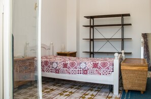 A bedroom with a bed, a wooden nightstand, a small table, a shelf unit, and a wicker chest of drawers. - The Sicilian Farmhouse (Palmeri - Ferrante)
