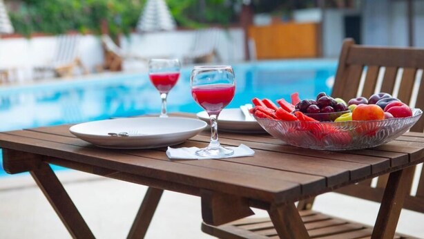 Ein Holztisch mit einer Obstschale und zwei Gläsern roter Flüssigkeit in der Nähe eines Schwimmbeckens.