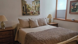 A bedroom with a large bed, two bedside tables, a window with blinds, and two wall-mounted lamps.