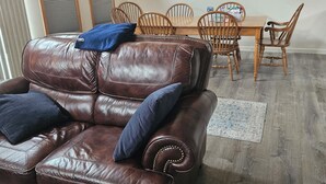 A brown leather sofa with two cushions, a wooden dining table with chairs, and a chandelier.