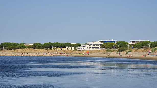 A beach with people sunbathing and swimming, a clear sky, and buildings in the background. - Près du Rivage Méridien (Le Grau-du-Roi)