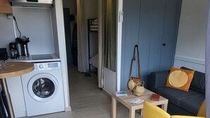 A compact living space with a washing machine, a small table, a sofa, and a door leading to another room. - Près du Rivage Méridien (Le Grau-du-Roi)
