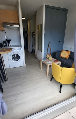 A compact living space with a washing machine, a small table, a sofa, and a door leading to another room.