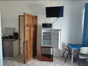 Ein kompakter Raum mit Holztür, einer kleinen Küchenzeile, einem weißen Kühlschrank, einer Mikrowelle, einem an der Wand montierten Fernseher, einem blauen Tisch und einem Stuhl.