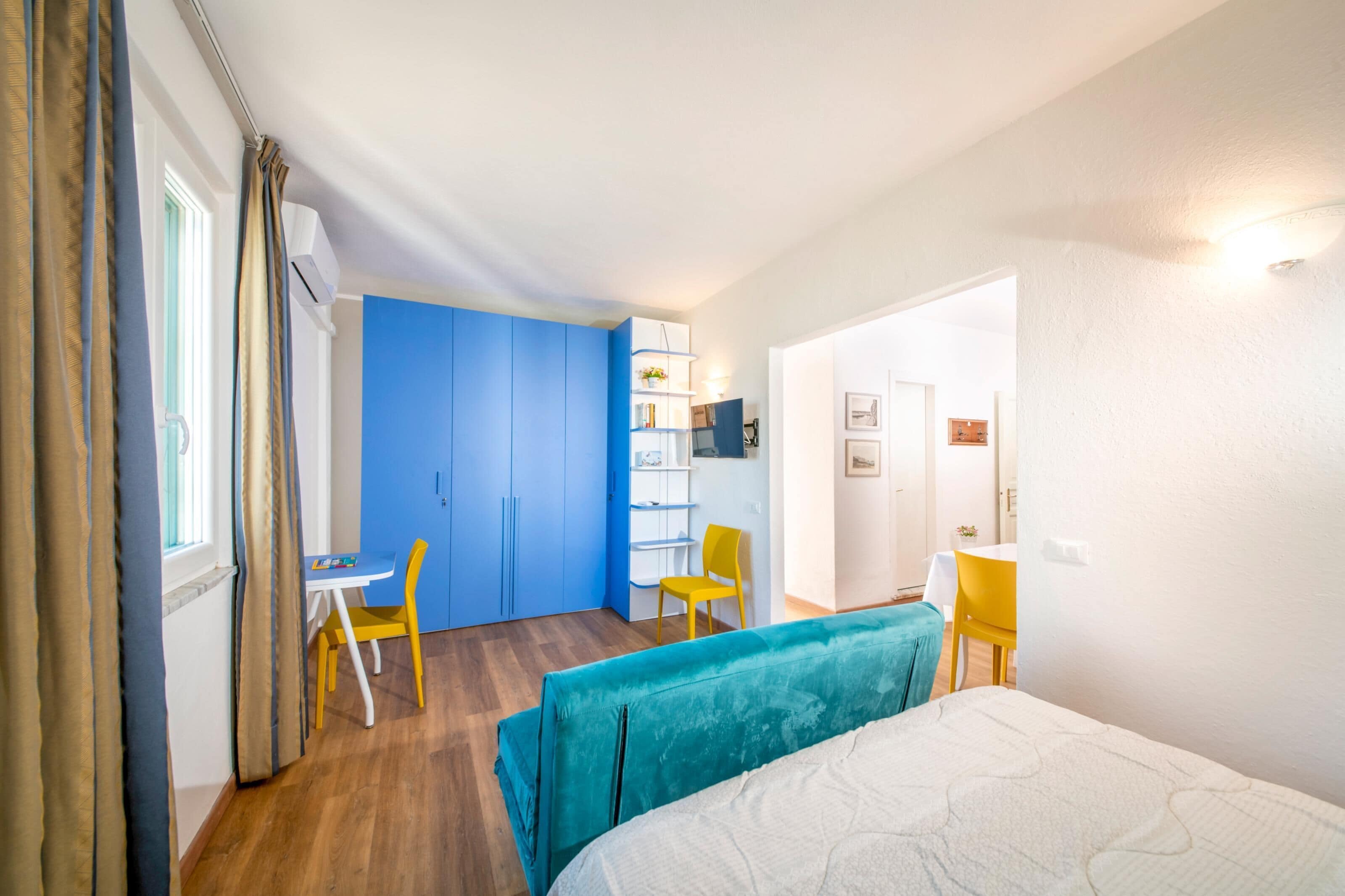 A room with a bed, a desk, and a chair. There is a blue wardrobe and a white wall with a shelf.